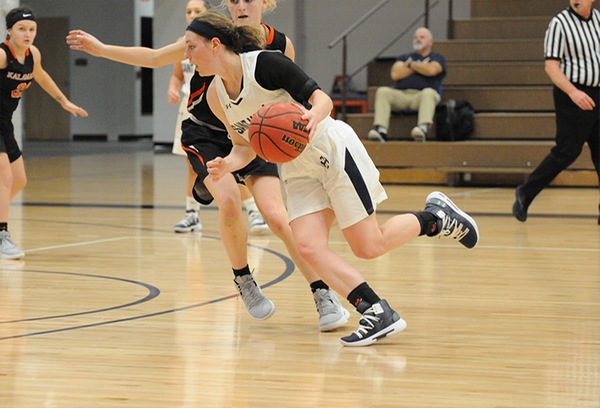 Erin Maloney, wearing a white uniform, dribbles the basketball past a defending player in a black uniform as two other players are visible in the background.