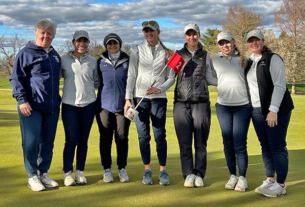 The team members and coach pose for a photo near a hole while holding a red golf flag with a white two.