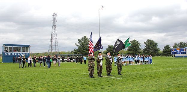 Four veterans present the colors prior to the start of a lacrosse game with the team, wearing light blue, visible in the background. A band of a dozen people play the National Anthem on the left side of the image.