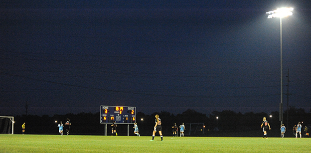 Several soccer players are visible at the bottom of the image during a night game at the Purcell Athletic Fields soccer field. A tall light is visible at the right of the image.