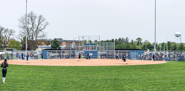 View of the softball field from the outfield looking in toward home plate. Several players are visible, and there are many people in the bleachers on both sides.