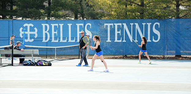 A tennis player is in the center of the photo wearing a black tank top and blue uniform bottom. A handful of other people are visible in the background as is the text "Belles Tennis" on a blue screen hanging on the fence.