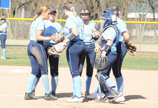 Six softball players wearing light blue tops and navy pants huddle up prior to the start of an inning.